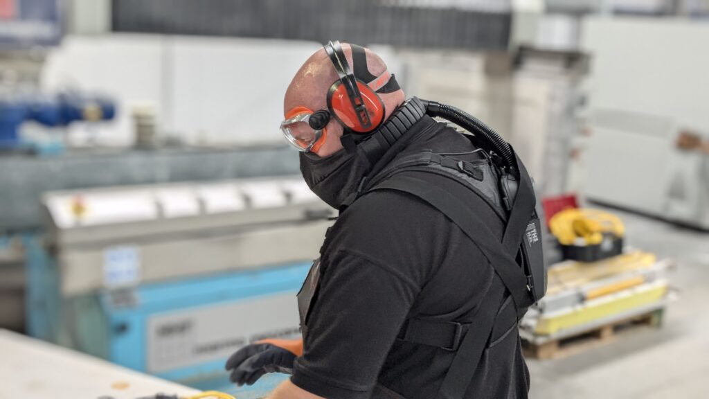 Stone fabricator wearing Optrel Swiss Air respirator, safety goggles and ear defenders in stone fabrication workshop